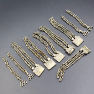 Pode incluir: Uma variedade de pe&ccedil;as de joalharia em metal prateado, incluindo correntes e elementos decorativos. As pe&ccedil;as variam em comprimento e design, algumas com extremidades quadradas ou circulares. As joias est&atilde;o dispostas sobre uma superf&iacute;cie cinza escuro.