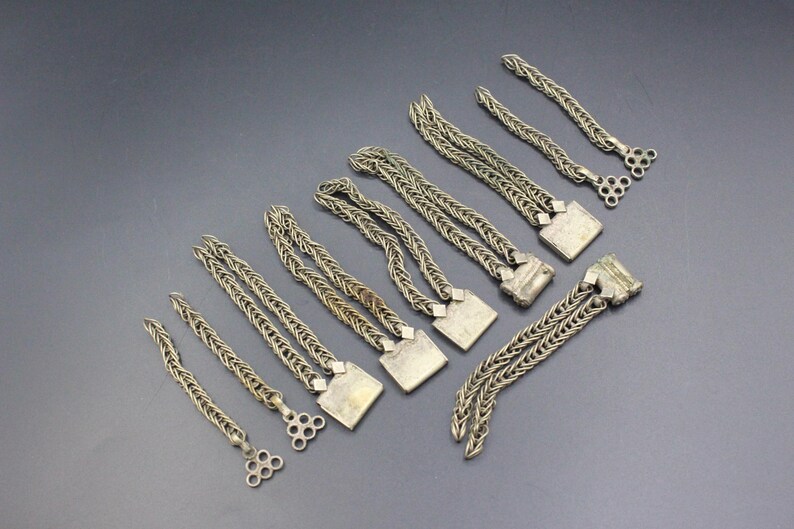 Pode incluir: Uma variedade de pe&ccedil;as de joalharia em metal prateado, incluindo correntes e elementos decorativos. As pe&ccedil;as variam em comprimento e design, algumas com extremidades quadradas ou circulares. As joias est&atilde;o dispostas sobre uma superf&iacute;cie cinza escuro.