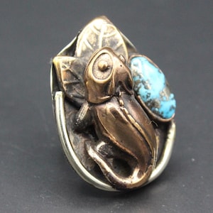 Puede incluir: Anillo de rana en tono bronce con una piedra turquesa. La rana está posada sobre una base en forma de hoja con detalles. El anillo tiene una banda plateada. Fondo oscuro.