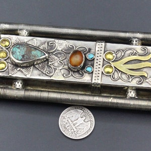 Puede incluir: Pieza decorativa de metal plateado ornamentada con piedras de color turquesa y &aacute;mbar, y detalles dorados. El objeto rectangular tiene un dise&ntilde;o detallado con una moneda para comparar el tama&ntilde;o. La pieza tiene dos barras paralelas y estriadas.