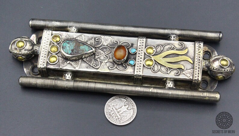 Puede incluir: Pieza decorativa de metal plateado ornamentada con piedras de color turquesa y &aacute;mbar, y detalles dorados. El objeto rectangular tiene un dise&ntilde;o detallado con una moneda para comparar el tama&ntilde;o. La pieza tiene dos barras paralelas y estriadas.