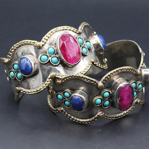 May include: Two silver cuff bracelets with intricate detailing. Each bracelet is adorned with a large, oval, magenta gemstone, complemented by smaller turquoise and blue stones. The bracelets have a vintage appearance.