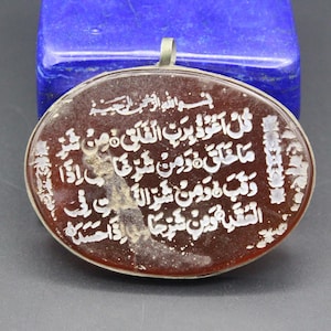 Könnte beinhalten: Ovaler Anhänger mit arabischer Schrift in Weiß auf einer rotbraunen Oberfläche, umrahmt von silberfarbenem Metall. Der Anhänger hat oben eine Schlaufe zum Aufhängen. Im Hintergrund befindet sich ein blauer Stein.