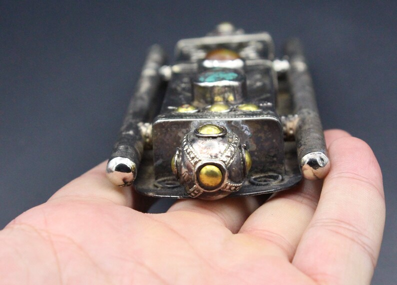 Puede incluir: Un colgante de metal plateado con un cuerpo rectangular y dos barras largas y paralelas. El colgante est&aacute; adornado con piedras de color turquesa y &aacute;mbar, y detalles dorados. El dise&ntilde;o tiene una est&eacute;tica vintage o antigua.