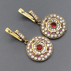 May include: A pair of gold-toned earrings with a circular design. Each earring features a central red stone surrounded by white and pink gemstones. The earrings have a lever back closure.