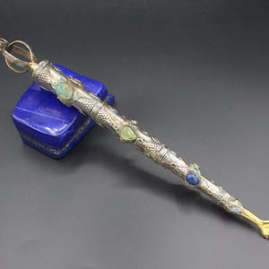 May include: An ornate silver and gold wand with inlaid gemstones, including turquoise and lapis lazuli. The wand features a detailed design and ends with a small hand. The wand is resting on a blue square object.