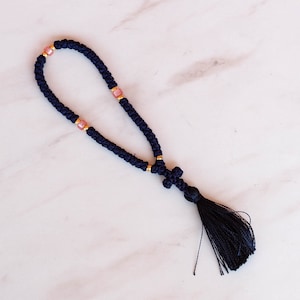 Puede incluir: Un cordón de oración negro anudado con cuentas rosadas y un borla. La cuerda está atada en un bucle y tiene una pequeña cruz en el extremo.