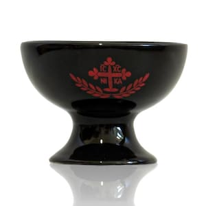 May include: A black ceramic chalice with a glossy finish. The chalice features a red emblem with text and a cross design. The chalice has a wide bowl, a pedestal base, and a smooth, reflective surface.