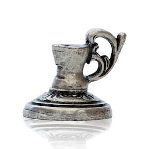May include: A small, silver-coloured metal candle holder featuring a circular base with embossed detailing and a single curved handle. The surface has a weathered, antiqued finish with dark highlights in the crevices of the metal structure.