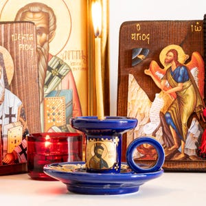 May include: A deep blue ceramic candle holder with a circular handle and a central iconographic portrait, holding a thin yellow beeswax candle. The holder is positioned in front of two wooden religious icons and a small red glass votive candle holder.