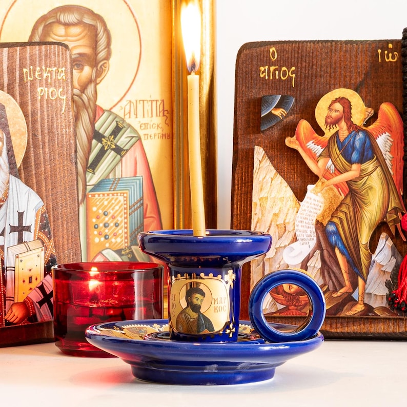 May include: A deep blue ceramic candle holder with a circular handle and a central iconographic portrait, holding a thin yellow beeswax candle. The holder is positioned in front of two wooden religious icons and a small red glass votive candle holder.
