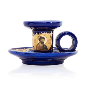 May include: A deep blue ceramic candle holder featuring a circular handle and a wide base. The central cylindrical section displays a gold-toned icon of a bearded figure with the Cyrillic text "АГЮС МАРКОС" printed on the front surface.
