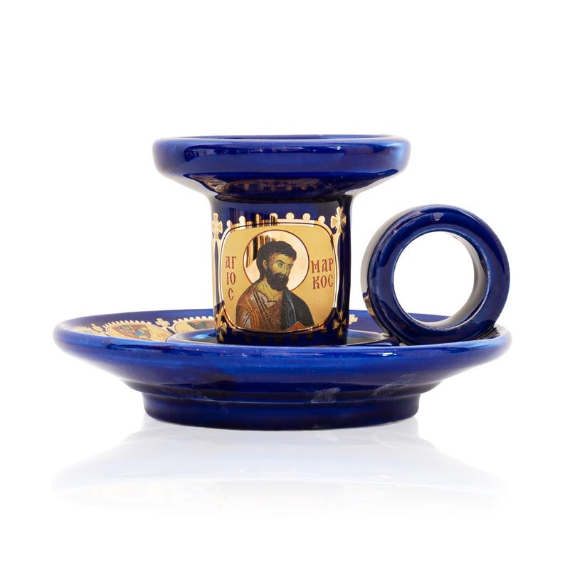 May include: A deep blue ceramic candle holder featuring a circular handle and a wide base. The central cylindrical section displays a gold-toned icon of a bearded figure with the Cyrillic text "АГЮС МАРКОС" printed on the front surface.
