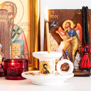 May include: A white ceramic candle holder with a circular handle and a central image of a bearded figure, holding a lit thin yellow candle. The holder sits before two wooden religious icons and a small red glass votive candle holder with a flame.