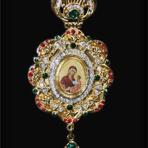 May include: Ornate gold-colored pendant featuring a central oval image of a religious figure with a child. The pendant is adorned with red, green, and clear gemstones, and has a crown-like top and a teardrop-shaped bottom element. The background is black.