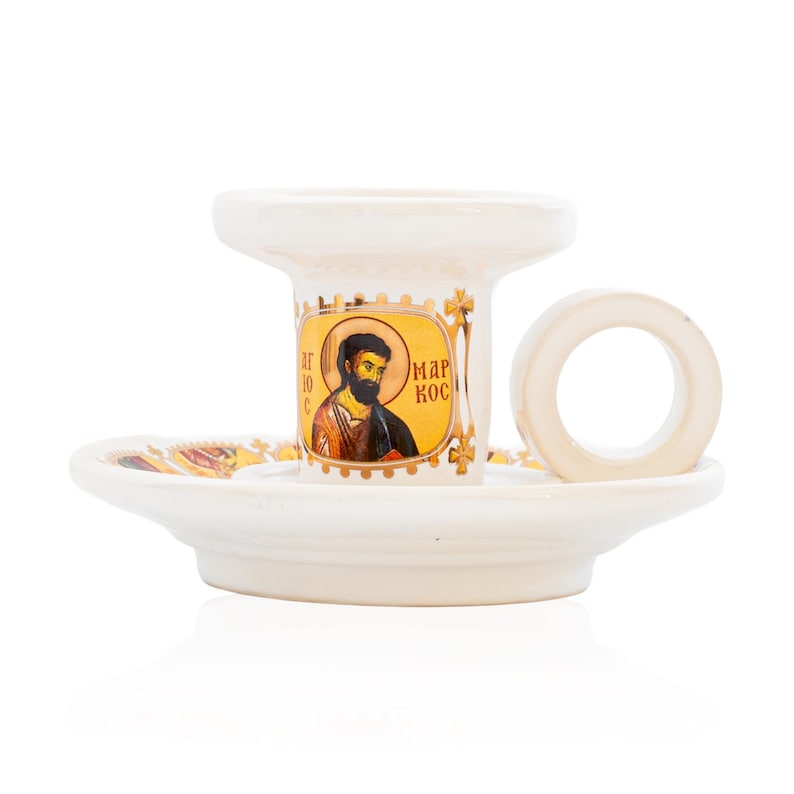 May include: A white ceramic candle holder with a circular handle and a wide base. The central column features a gold-toned icon of a bearded figure with the Greek text "&Alpha;&Gamma;&Iota;&Omicron;&Sigma; &Mu;&Alpha;&Rho;&Kappa;&Omicron;&Sigma;" printed on the front. The base is decorated with gold accents.