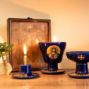Può includere: Una natura morta con oggetti religiosi. Tre candelabri blu in ceramica con dettagli dorati e candele accese. Un'icona di Gesù è su un portacandele. Sono presenti anche un piccolo vaso con una pianta e un'icona incorniciata.
