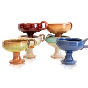 Pode incluir: Um conjunto de seis taças de cerâmica com pedestal em várias cores, incluindo vermelho, bege, verde, azul e bege. Cada taça tem uma alça e uma forma de tigela arredondada, ideal para servir bebidas ou sobremesas.