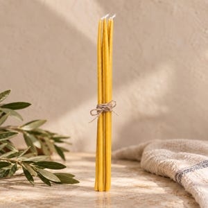 Puede incluir: Un manojo de cuatro velas cónicas altas, delgadas y de color amarillo dorado, atadas con un trozo de cuerda marrón natural. Las velas tienen una forma cilíndrica lisa con una parte superior puntiaguda y una mecha blanca visible en cada punta.