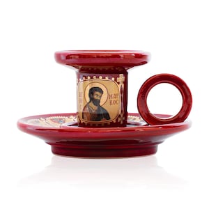 May include: A glossy red ceramic candle holder featuring a circular handle and a central pillar decorated with a gold-toned icon of a bearded figure. The text "&Alpha;&Gamma;&Iota;&Omicron;&Sigma; &Mu;&Alpha;&Rho;&Kappa;&Omicron;&Sigma;" is printed in gold on the pillar, which sits on a wide, matching red base.