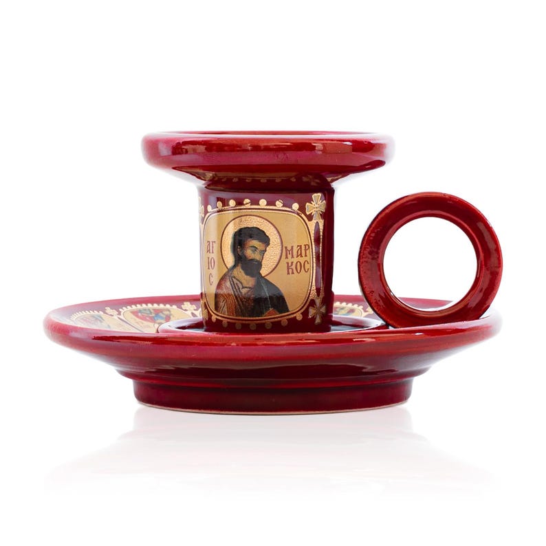 May include: A glossy red ceramic candle holder featuring a circular handle and a central pillar decorated with a gold-toned icon of a bearded figure. The text "&Alpha;&Gamma;&Iota;&Omicron;&Sigma; &Mu;&Alpha;&Rho;&Kappa;&Omicron;&Sigma;" is printed in gold on the pillar, which sits on a wide, matching red base.