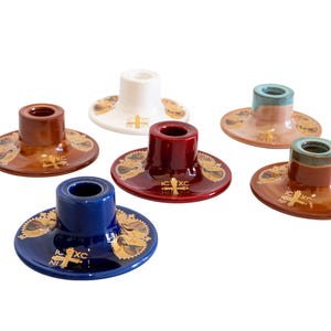 May include: A collection of six ceramic candle holders in various colors including white, blue, red, and brown. Each holder features a wide circular base with gold-colored religious iconography and the text IC XC NI KA printed on the surface.