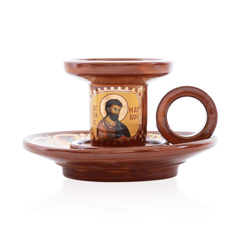 May include: A brown ceramic candle holder with a wide base and a circular side handle. The central column features a gold-toned icon of a bearded figure with the Greek text "&Alpha;&Gamma;&Iota;&Omicron;&Sigma; &Mu;&Alpha;&Rho;&Kappa;&Omicron;&Sigma;" printed on the surface, surrounded by decorative gold accents.