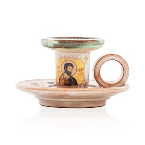 May include: A ceramic candle holder with a beige and brown glaze, featuring a circular handle and a central cylindrical pillar. The pillar displays a religious icon of a bearded figure with the Greek text "&Alpha;&Gamma;&Iota;&Omicron;&Sigma; &Mu;&Alpha;&Rho;&Kappa;&Omicron;&Sigma;" printed on the front.