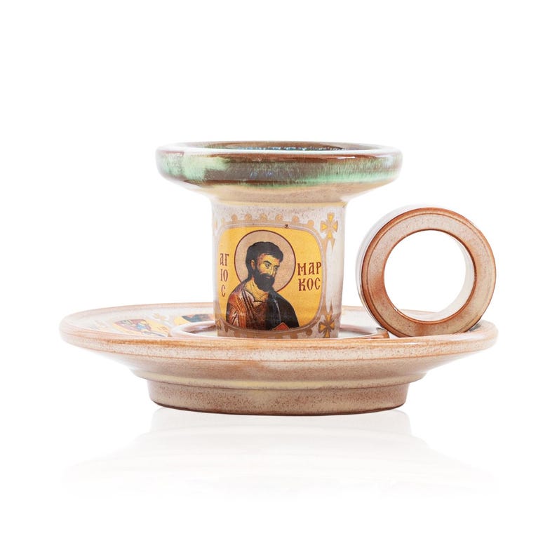 May include: A ceramic candle holder with a beige and brown glaze, featuring a circular handle and a central cylindrical pillar. The pillar displays a religious icon of a bearded figure with the Greek text "&Alpha;&Gamma;&Iota;&Omicron;&Sigma; &Mu;&Alpha;&Rho;&Kappa;&Omicron;&Sigma;" printed on the front.
