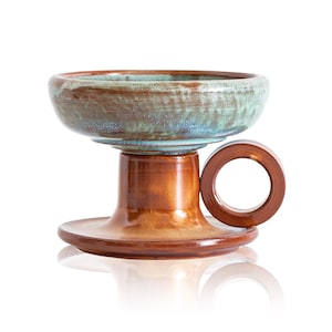 May include: A ceramic pedestal bowl with a brown and teal glaze. The bowl features a circular handle and a wide base. The interior of the bowl has a teal and blue colour, while the exterior is brown.