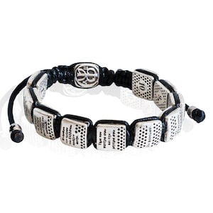 Könnte beinhalten: Silberfarbenes Armband mit quadratischen Anhängern und schwarzem Kordel. Jeder Anhänger hat ein gepunktetes Muster und griechischen Text. Das Armband hat einen zentralen Anhänger mit Logo und verstellbaren Knoten.