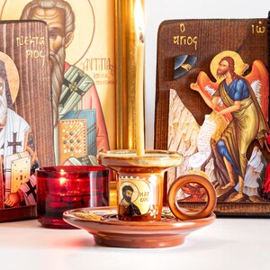 May include: A ceramic candle holder with a brown glaze and a circular handle, featuring a small icon of Saint Mark with the text "MAPKOC". A thin, light-colored beeswax candle is lit inside the holder, positioned in front of religious icons.