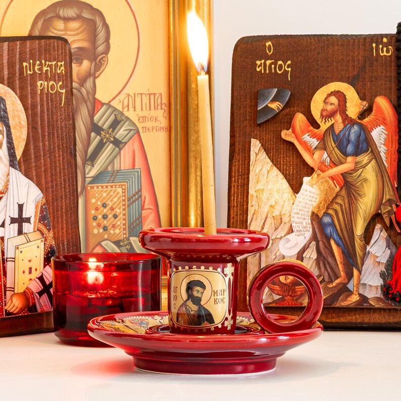 May include: A red ceramic candle holder with a circular handle and a central icon of a bearded figure labeled with the Greek text "&Alpha;&Gamma;&Iota;&Omicron;&Sigma; &Mu;&Alpha;&Rho;&Kappa;&Omicron;&Sigma;". A thin, lit beeswax candle stands in the center, positioned in front of two wooden religious icons.