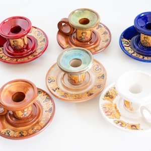 May include: A collection of six ceramic candle holders in red, brown, blue, beige, and white. Each holder features a circular base decorated with small religious icon portraits and a central pillar with a handle and a top opening for a candle.