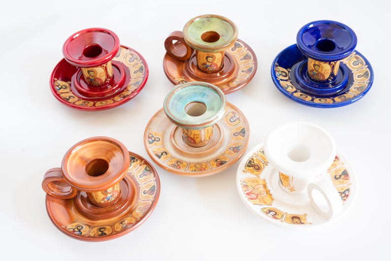 May include: A collection of six ceramic candle holders in red, brown, blue, beige, and white. Each holder features a circular base decorated with small religious icon portraits and a central pillar with a handle and a top opening for a candle.