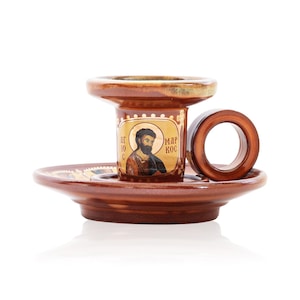 May include: A brown glazed ceramic candle holder with a circular handle and a wide base. The central column features a gold-toned icon of a bearded figure with the Greek text "&Alpha;&Gamma;&Iota;&Omicron;&Sigma; &Mu;&Alpha;&Rho;&Kappa;&Omicron;&Sigma;" printed on either side of the portrait in black lettering.