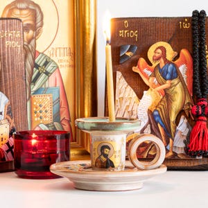 May include: A ceramic candle holder with a light beige finish and a green rim, featuring a circular handle and a central icon of a bearded figure with the text "AGIOS MARKOS". A thin yellow candle burns inside, set against a backdrop of religious icons.