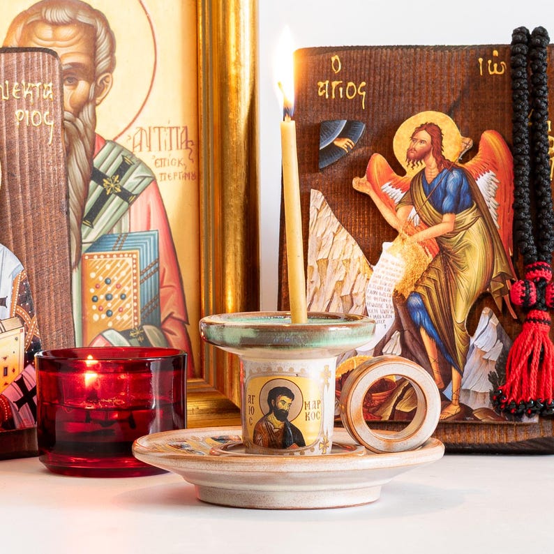 May include: A ceramic candle holder with a light beige finish and a green rim, featuring a circular handle and a central icon of a bearded figure with the text "AGIOS MARKOS". A thin yellow candle burns inside, set against a backdrop of religious icons.
