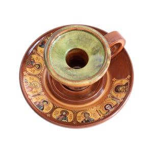 May include: A brown ceramic candle holder with a handle and a wide saucer base. The saucer features a circular border of small, colorful religious icon portraits with Greek text. The top of the candle holder has a smooth, light green glazed finish.