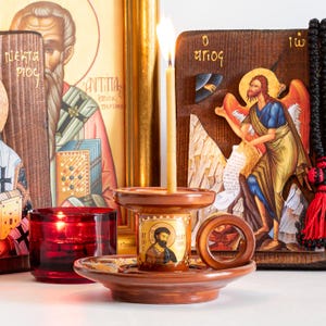 May include: A brown ceramic candle holder with a circular handle and a central icon of a bearded figure labeled with Greek text. A thin lit candle stands in the center. Beside it sits a red glass votive holder and two wooden religious icons.