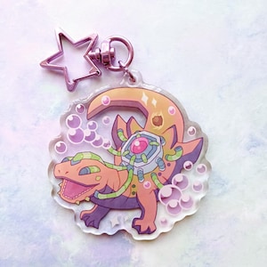 May include: A clear acrylic keychain featuring a cartoon illustration of a pink and orange lizard with a mechanical device on its back. The lizard is surrounded by purple and white bubbles.