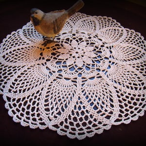 Puede incluir: Un delicado encaje de ganchillo blanco con un diseño circular. El encaje tiene un centro elevado con un patrón floral y un borde festoneado. Una pequeña figurita de pájaro se coloca encima del encaje.