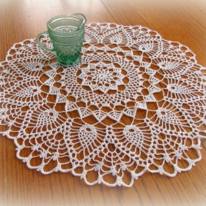 May include: A white crocheted doily with a delicate lace pattern, featuring a circular design with multiple layers. The doily is placed on a wooden table with a green glass pitcher.