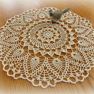 以下が含まれることがあります： 繊細な花柄のベージュのクロシェットドイリー。ドイリーは円形で、中央に小さな鳥の置物があります。