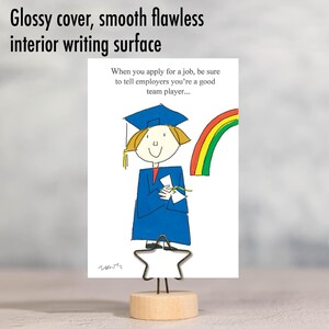 May include: A white card with a cartoon illustration of a graduate holding a diploma and a rainbow in the background. The card is displayed on a wooden stand with a star-shaped metal clip. The text on the card reads: "When you apply for a job, be sure to tell employers you're a good team player..."