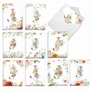 May include: A collection of white greeting cards and envelopes featuring the letter J decorated with various watercolor floral arrangements in shades of pink, orange, blue, green, and yellow, displayed in multiple stacks.