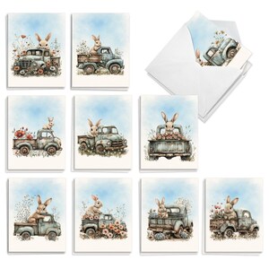 May include: A collection of greeting cards featuring watercolor illustrations of light brown rabbits sitting in the beds of vintage blue pickup trucks surrounded by various wildflowers against a soft blue sky background, with one white envelope.