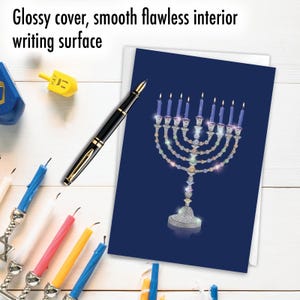May include: A greeting card with a blue background features a jeweled menorah with lit candles. A black pen with gold accents rests on the card. The text "Glossy cover, smooth flawless interior writing surface" is at the top.