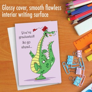 May include: A graduation card with a cartoon green dragon wearing a graduation cap. The card has a purple background and the text "You've graduated! So go ahead..."