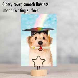 May include: A graduation card featuring a dog wearing a graduation cap and holding a diploma in its mouth. The card has a blue background with a rainbow in the top corner.